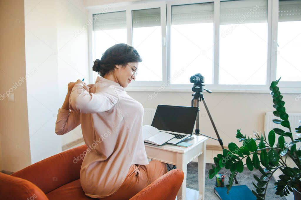 Tired caucasian woman with eyeglasses having online lessons using a camera and laptop while stretching. Home network. Online education. Online video.