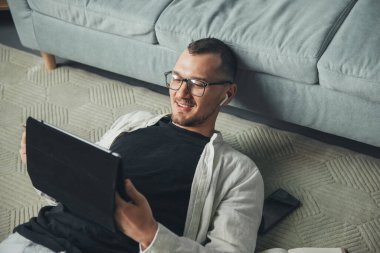 Tablet kullanan bir adam yerde yatarken ve başını kanepeye yaslarken. Ön manzara. Çevrimiçi çalışıyor