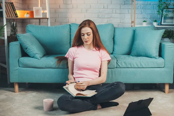 La mujer de negocios escribe en un cuaderno, sentada en el suelo, apoyada en el sofá. Educación a distancia en línea y trabajo. Pelo rojo. Cara pecosa. — Foto de Stock