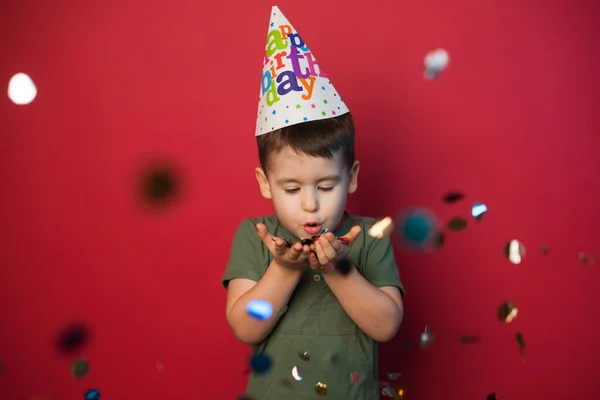Vit pojke i en festlig mössa blåser konfetti till kameran på en röd bakgrund. Begreppet en rolig och bullriga barns semester. — Stockfoto