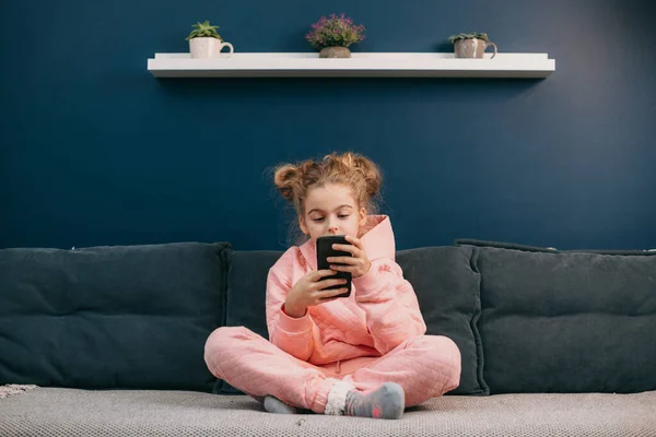 Přední pohled na malou blondýnku sedící doma na pohovce s mobilním telefonem. Děti a přístroje. — Stock fotografie
