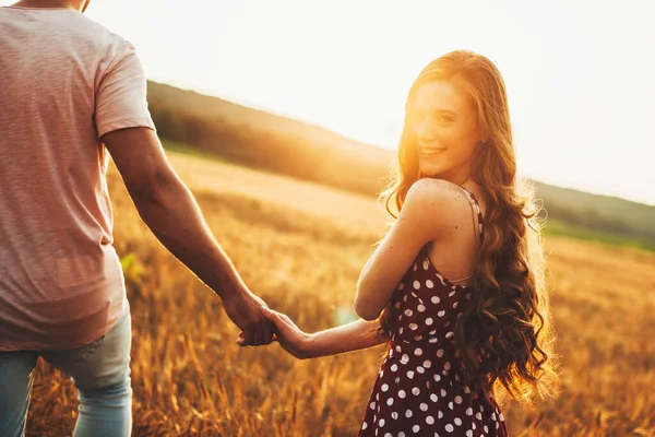 Una mujer y un hombre caminando por el campo tomados de la mano en un día soleado. Mujer pelirroja sonriendo a la cámara. Vacaciones de verano. Campo de trigo. —  Fotos de Stock