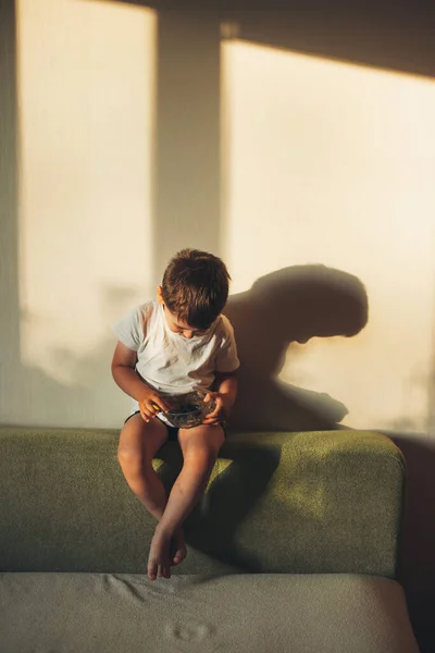 Niño sentado en el sofá en casa comiendo arándanos. Comida saludable. —  Fotos de Stock