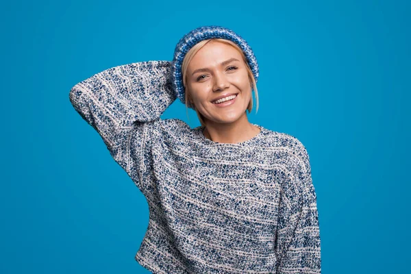 Mujer rubia sonriente con suéter de punto y sombrero posando sobre fondo de estudio azul aislado. Mujer bastante joven. — Foto de Stock