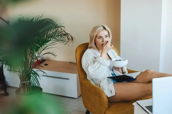 Blonde caucasian woman sitting in armchair and looking away holding a notebook on his legs while eating cereals. Morning lifestyle. Distance education. Healthy — Stockfoto