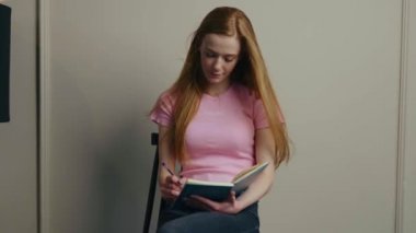 Caucasian woman sitting on the chair at home and writing in her planner book. Red-haired woman. Study day concept. Writing education concept. Lifestyle concept