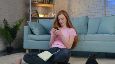 Woman with copybook in hand sitting at floor using cellphone to send a message. Distance education. Holding hands.
