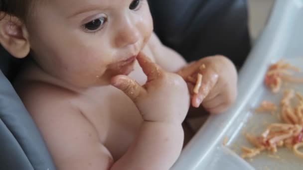 Messy baby girl use hand eating spaghetti sitting in high chair at home. Close-up portrait. Baby care. — Video