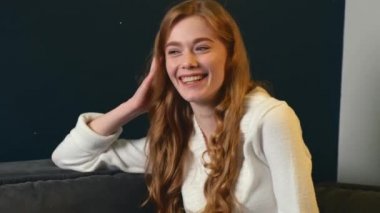 Adorable woman with ginger hair and freckles is sitting on the couch and smile at camera