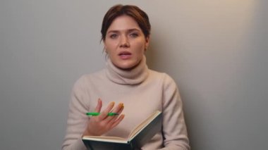 Short haired woman is speaking in front of camera while holding a book and pen on a gray studio wall