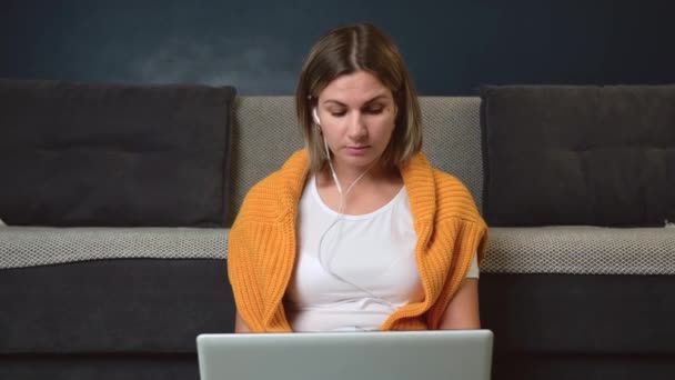 Caucasian woman is sitting on the floor with a laptop using earphones during video meeting — Stockvideo