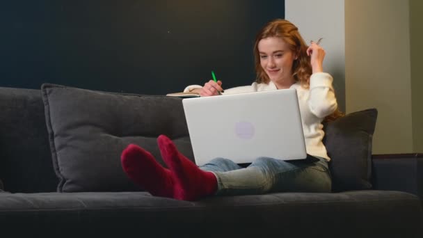 白人の生姜の女性はソファに横たわっている間、トピックを書いているとオンラインレッスンでラップトップを使用しています — ストック動画