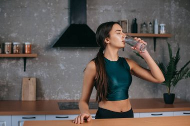 Caucasian woman standing in the home kitchen drinking fresh water after doing workout. Dieting, healthy lifestyle, weight loss concept. Beauty concept. Home