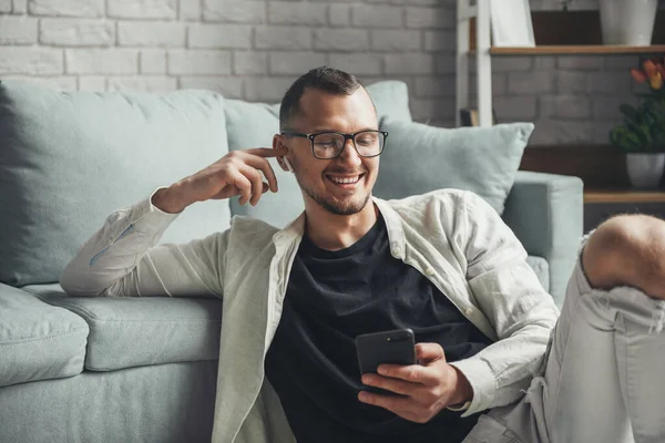 Portrait of a smiling man using cellphone and wireless earphones while sitting on floor at home. — Fotografia de Stock