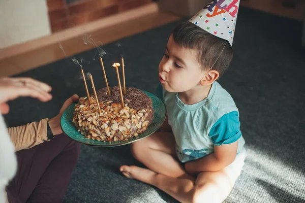 Boy blowing burning candles on his birthday cake given by moms hands while sitting on the floor during his birthday party. — Zdjęcie stockowe