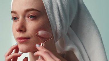 Close-up portrait of a woman with towel on head using jade roller on face. Rejuvenation treatment. Facial skin treatment. Facial skincare.
