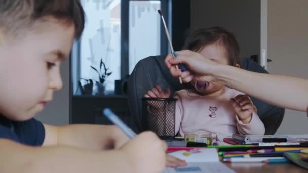 Frontansicht eines kleinen Mädchens, das sich auf das Zeichnen mit Farbstift konzentriert. Farbstifte liegen auf einem Tisch. Bruder macht Hausaufgaben. Mutter hilft. — Stockvideo