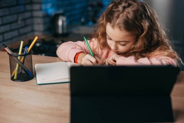Little girl studying and taking notes using a digital tablet while sitting at home.