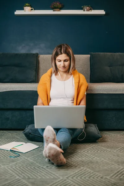 Kulaklıklı beyaz bir kadın dizüstü bilgisayar ekranına bakıyor. Uzaktan kurs alırken ya da evde dizüstü bilgisayar başında yerde oturuyor. Ön manzara. İnternette okuyan kız — Stok fotoğraf