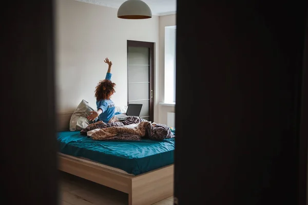 Curly woman in pajamas sitting on bed in front of laptop and watching online lesson while making notes in copybook. Internet technology. — Foto de Stock