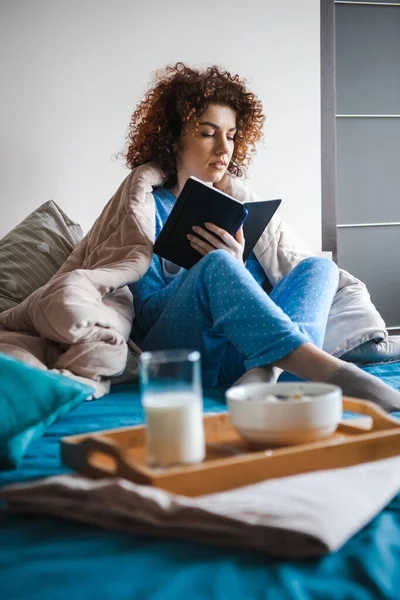 Curly woman with pen, book and notebook, writting somethning in bed, preparing for exam. Beautiful morning bed, great design for any purposes. — Fotografia de Stock