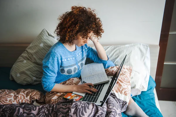 Curly brunette woman sitting in bed making notes in notepad using laptop. Bedtime. — Foto de Stock