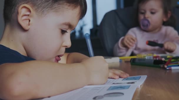 Video med en pojke i sitter vid bordet med sin lillasyster gör anteckningar i copybooks medan du gör läxor. Utbildning för barn. Distansutbildning. — Stockvideo