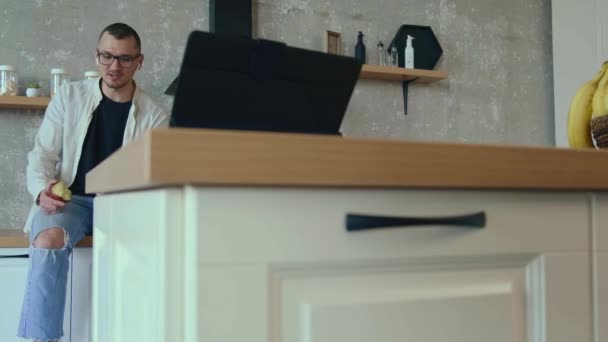 Caucasiano sorrindo homem falando ao telefone enquanto come uma maçã sentada na cozinha em casa. Pessoas a falar. Que casa inteligente. Comunicação móvel. — Vídeo de Stock