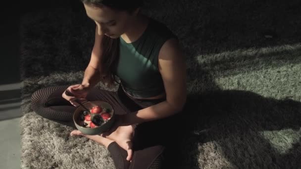 Βίντεο μιας γυναίκας που κάθεται στο πάτωμα στο σπίτι, χαλαρώνοντας και τρώγοντας ένα μπολ με γκρανόλα ή κυλιόμενη βρώμη με κράνμπερι. Φυσική βιολογική διατροφή. — Αρχείο Βίντεο