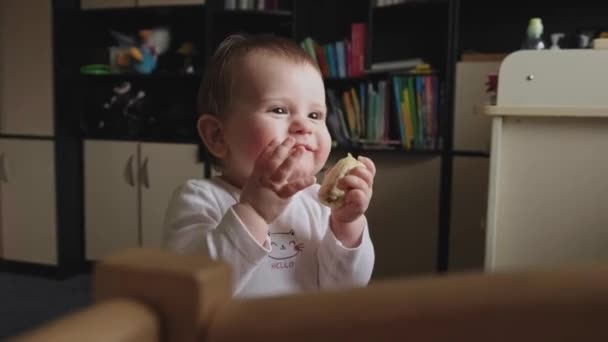 Video med en glad liten flicka som sitter på golvet och äter banan med aptit. Närbild porträtt. Vacker flicka. Familjeomsorg. Barnomsorg. — Stockvideo