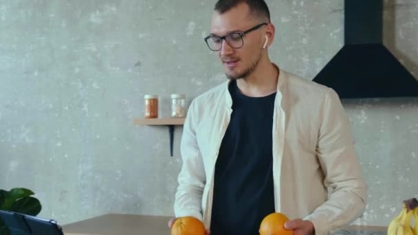 Man standing in the kitchen and attending an online conference while juggling with two oranges. Modern portrait. Internet technology. — Stock Video