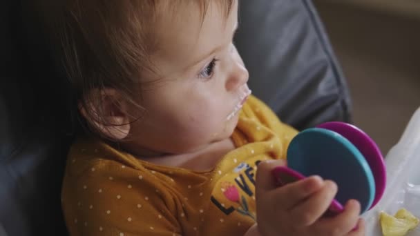 Video van een meisje dat met haar speelgoed speelt terwijl ze eet, zittend aan de kindertafel. De moeders geven de baby water om uit de fles te drinken.. — Stockvideo