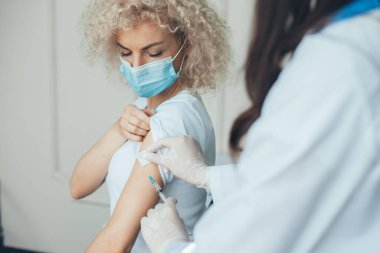 Tıbbi maskeli kıvırcık kadın bir hemşireden bir doz koronavirüs aşısı alıyor. Coronavirus aşısı. Grip salgını tıbbi sağlık. Salgın
