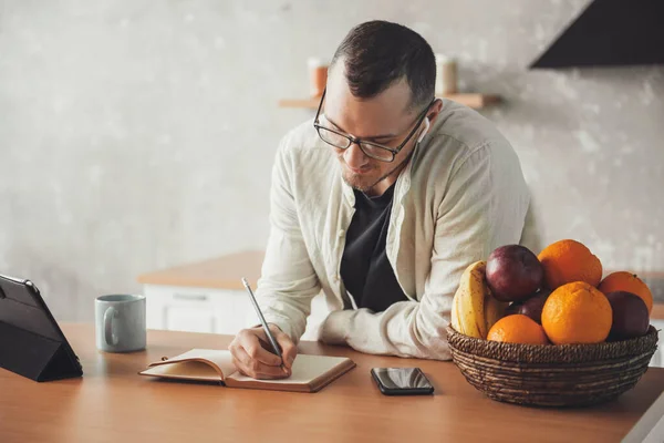 Mannen sitter vid bordet i köket och skriver ner dagboken med prylar när han studerar hemifrån. Fjärrarbete. — Stockfoto