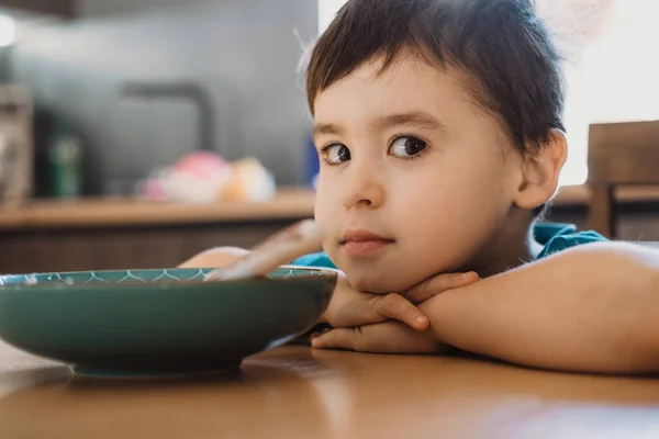Мальчик, сидящий за кухонным столом и наклоняющий голову от стола, смотрит в камеру с миской. — стоковое фото