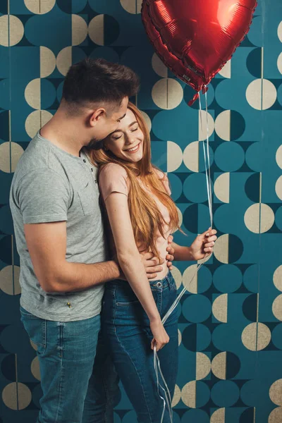 青い壁の背景に隔離された心の風船でポーズをしながら笑顔のカップル立っている。バレンタインデー。美しい女の子. — ストック写真