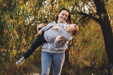 Anne, parkta ilginç bir oyun oynarken oğlunu kucağında havaya kaldırdı. Havalı anne oğlu dışarıda gülümsüyor, her şey için harika bir tasarım.