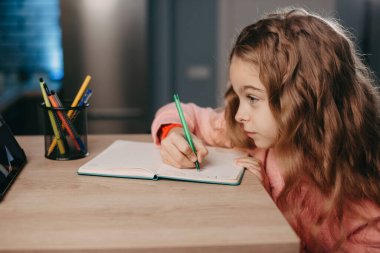 Kız not defterine yazarken masada oturup tabletle online kurs izliyor. Çevrimiçi öğrenim okulu. Online öğretim.