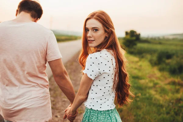 Vue arrière d'un couple se tenant la main et marchant sur une route de campagne. Fille aux cheveux roux et taches de rousseur tournant la tête vers la caméra. Vacances d'été. — Photo