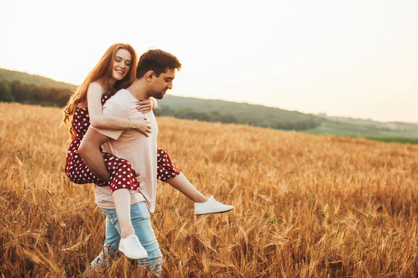 Man carrying his red-haired girlfriend on his back as he walks through a wheat field. Piggy back — Stock Photo, Image