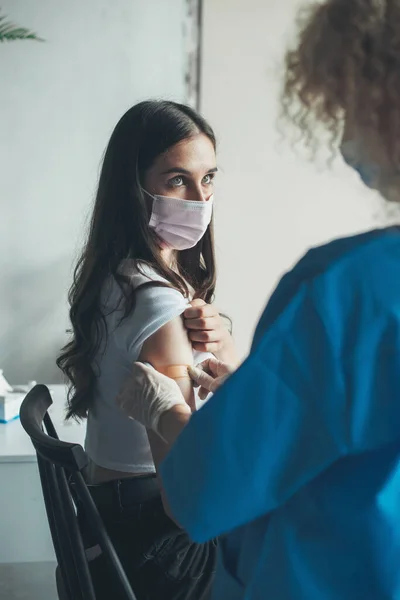 Pandangan belakang seorang perawat menerapkan plester perekat pada lengan seorang wanita setelah menerima vaksin. Konsep kesehatan medis. Konsep vaksinasi orang. Medis — Stok Foto