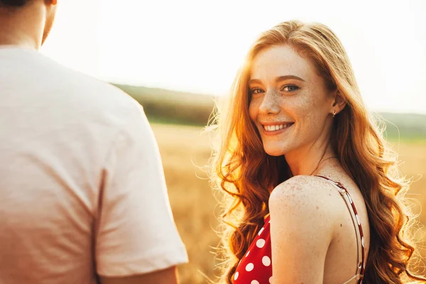 Close-up portret van een roodharige vrouw die haar hoofd naar de camera draait terwijl ze met haar vriendje in het tarweveld loopt. Verliefd stel — Stockfoto