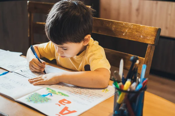 Pequeño niño caucásico utilizar la escritura a lápiz en el cuaderno sentado en la mesa de madera en casa. Aprendiendo de casa. Estudiar, hacer los deberes. —  Fotos de Stock
