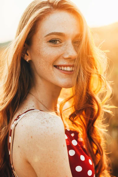 Közelkép egy vörös hajú, szeplős arcú romantikus nőről, aki élvezi a természetet, mosolyog és a kamerába néz. Napfény naplementekor. — Stock Fotó
