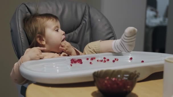 Bebê sentado em uma cadeira com as pernas estendidas e a mão na boca comendo uma romã. — Vídeo de Stock