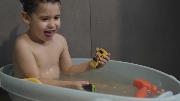 Niño caucásico bañándose con sus juguetes favoritos y salpicando agua con sus pies. Retrato para diseño sanitario. Sonriente niño feliz. — Vídeos de Stock