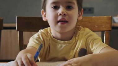 Küçük beyaz öğrenci, ahşap masaya konsantre yüzlü bir kitap yazmak için deftere kalem kullanıyor. Çocuk gelişimi. Çocuk eğitimi. Önde