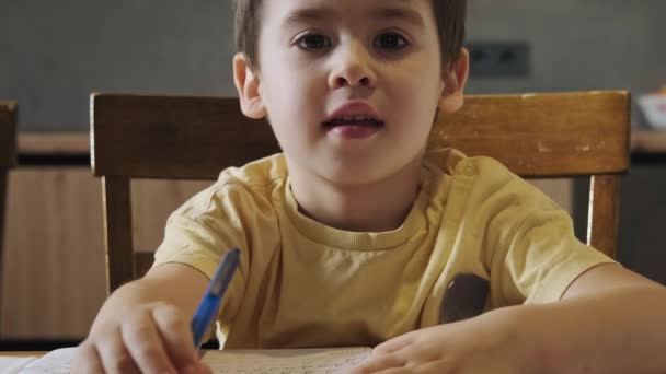 Ο μικρός καυκάσιος μαθητής χρησιμοποιεί μολύβι γράφοντας στο σημειωματάριο για να γράψει βιβλίο με συγκεντρωμένο πρόσωπο στο ξύλινο τραπέζι. Ανάπτυξη παιδιών. Παιδική εκπαίδευση. Εμπρός — Αρχείο Βίντεο