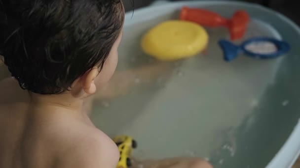 Widok z tyłu chłopca stojącego w wannie i bawiącego się dwoma zabawkowymi samochodami wrzucającego je do wody i opryskującego wodą. Uśmiechnięte dziecko. — Wideo stockowe