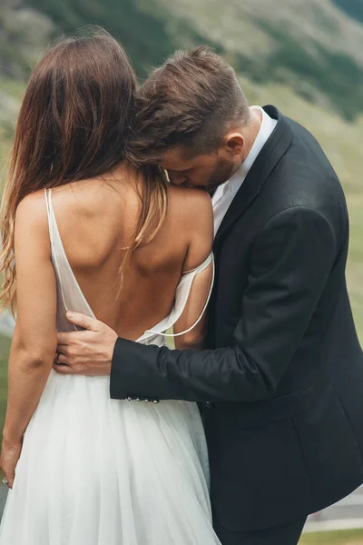Groom in a dark suit kisses a beautiful brunette bride on her necked shoulder. Lifestyle, female beauty concept. Family lifestyle concept.